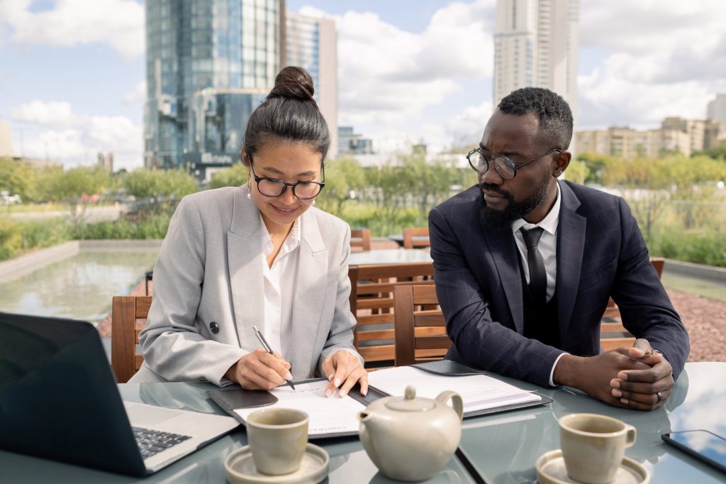 Two young elegant business partners sitting by table while one of them signing contract